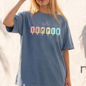 May include: A blue t-shirt with the words "hello SUMMER" printed in pastel colors. The shirt features a popsicle design. The person is holding a camera and wearing a white skirt. A straw bag with flowers is in the foreground.