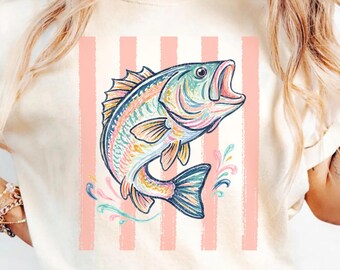 May include: An ivory short-sleeve t-shirt featuring a graphic print of a jumping fish in shades of blue, pink, and yellow. The design includes vertical light pink stripes behind the fish and colorful water splashes at the base of the illustration.