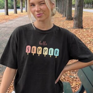 May include: Black t-shirt with the text "hello SUMMER" and popsicle graphics in pastel colors. The shirt is worn by a person sitting on a park bench. The shirt is a casual summer wear.