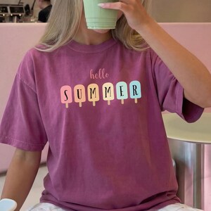 May include: A magenta t-shirt with the words "hello SUMMER" printed on the front. The word "SUMMER" is designed with colorful popsicle graphics. The person is holding a green drink in a clear cup.
