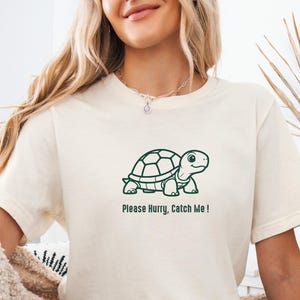 Peut inclure: Un t-shirt à manches courtes de couleur naturelle avec une illustration au trait vert foncé d'une tortue centrée sur la poitrine. Sous le graphique de la tortue, le texte indique "Please Hurry, Catch Me !" dans une police sans empattement.