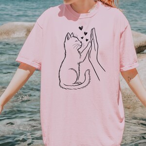 May include: A light pink t-shirt featuring a black line drawing of a cat high-fiving a hand, with three small hearts above them. The word "BLOSSOM" is printed in black at the bottom right corner of the shirt.