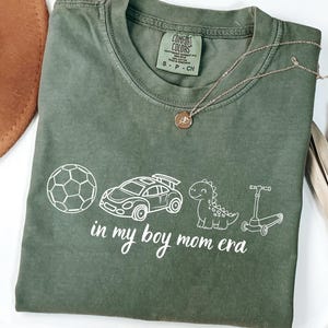 May include: A moss green t-shirt with the text "in my boy mom era" and white illustrations of a soccer ball, race car, dinosaur, and scooter. A gold necklace is draped across the shirt.