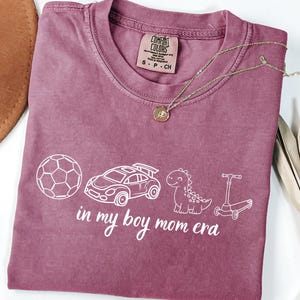 May include: A mauve Comfort Colors t-shirt with a white graphic of a soccer ball, race car, dinosaur, and scooter. The text "in my boy mom era" is below the graphics. A gold necklace is draped across the shirt.