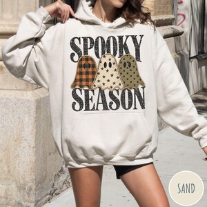 May include: A sand-colored hooded sweatshirt featuring a graphic print of three ghosts with different patterns: orange plaid, cream with black stars, and olive green with black dots. The text SPOOKY SEASON is printed in black above and below the ghosts.