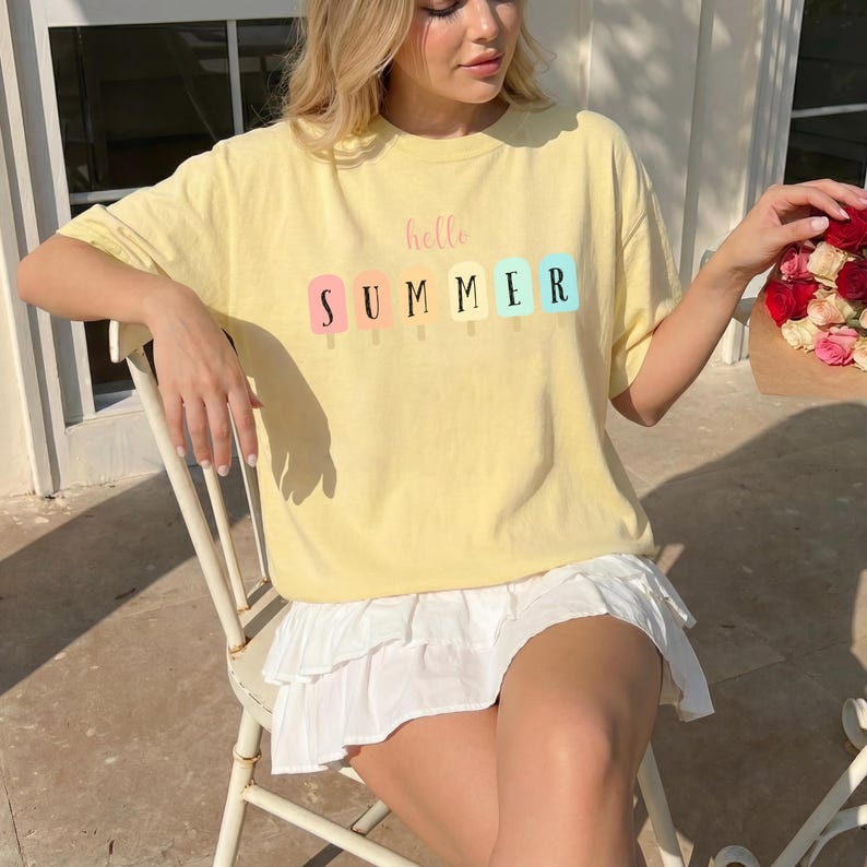 May include: A light yellow t-shirt with the words "hello SUMMER" printed on the front in pastel colors. The shirt is paired with a white skirt. The overall style is casual and summery.