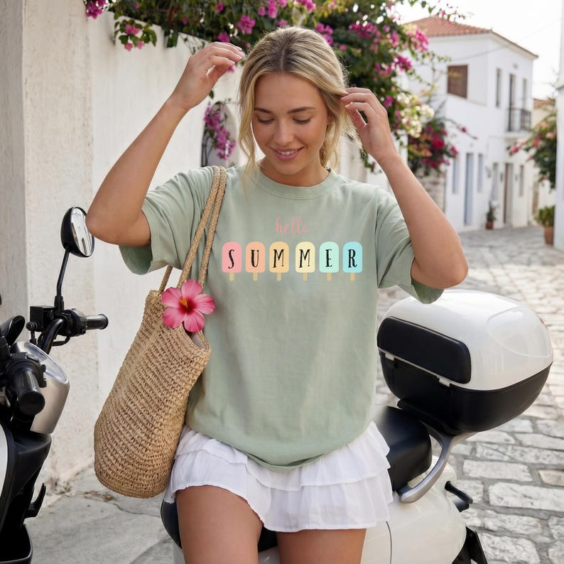 May include: A light green t-shirt with the words "hello SUMMER" printed on the front in pastel colors. The shirt is paired with a white skirt and a woven tote bag with a pink flower. The person is standing next to a scooter.