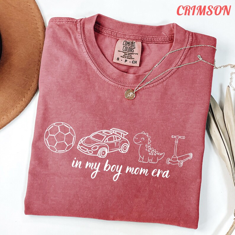 May include: Crimson-colored t-shirt with white graphic of a soccer ball, race car, dinosaur, and scooter. Text reads "in my boy mom era". The shirt has a crew neck and a Comfort Colors tag.