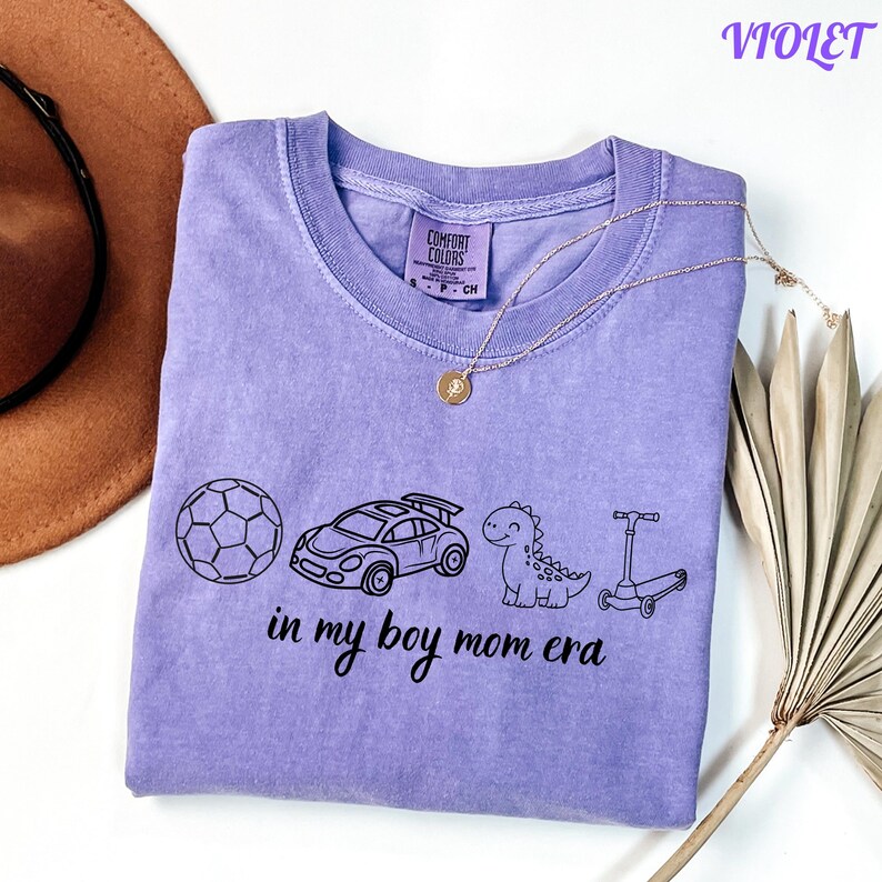 May include: A violet Comfort Colors t-shirt with a graphic of a soccer ball, race car, dinosaur, and scooter. The text "in my boy mom era" is printed below the graphics. A gold necklace is draped across the shirt.
