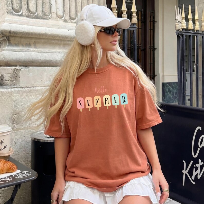 May include: Rust-colored t-shirt with the words "hello SUMMER" and colorful popsicle graphics. The shirt is a short-sleeved, casual style. The image shows a person wearing the shirt with a white hat and sunglasses.