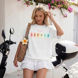 May include: White t-shirt with the text "hello SUMMER" above a row of colorful popsicle illustrations. The shirt is worn with a white skirt and a woven bag. The background includes a scooter and a wall with flowers.