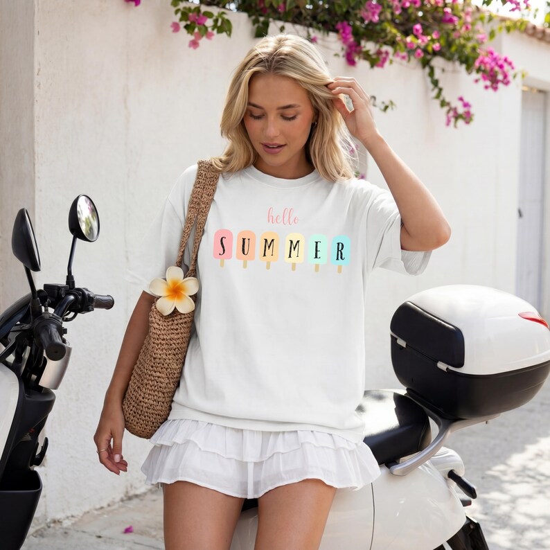 May include: White t-shirt with the text "hello SUMMER" above a row of colorful popsicle illustrations. The shirt is worn with a white skirt and a woven bag. The background includes a scooter and a wall with flowers.
