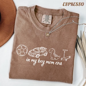 May include: Espresso-colored t-shirt with white graphic of a soccer ball, race car, dinosaur, and scooter. The text "in my boy mom era" is below the graphics. A silver necklace with a pendant is on the shirt.