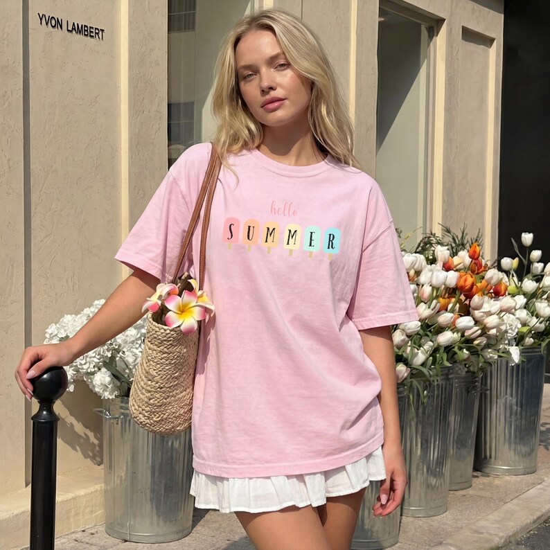 May include: A light pink t-shirt with the words "hello SUMMER" printed on the front, featuring colorful popsicle illustrations. The shirt is paired with a white skirt and a straw bag with flowers.