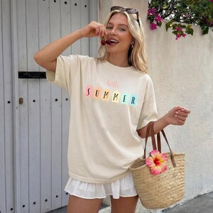 May include: Cream-colored t-shirt with the words "hello SUMMER" in pastel colors. The shirt is paired with a white skirt and a straw tote bag with a pink flower.