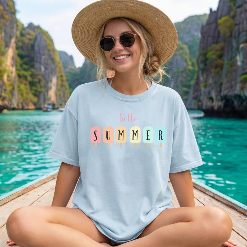 May include: Light blue t-shirt with the words "hello SUMMER" and popsicle graphics in pastel colors. The shirt is worn by a person wearing a straw hat and sunglasses, sitting on a boat.