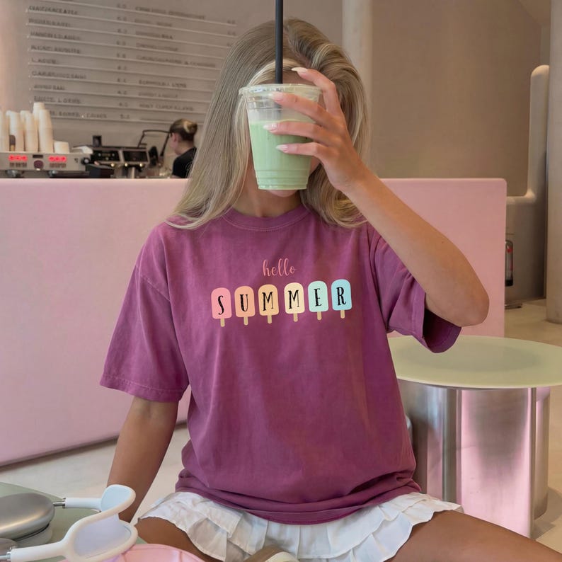May include: A magenta t-shirt with the words "hello SUMMER" printed on the front. The word "SUMMER" is designed with colorful popsicle graphics. The person is holding a green drink in a clear cup.