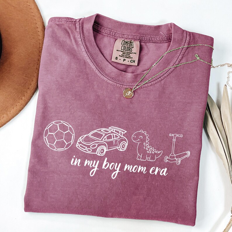 May include: A mauve Comfort Colors t-shirt with a white graphic of a soccer ball, race car, dinosaur, and scooter. The text "in my boy mom era" is below the graphics. A gold necklace is draped across the shirt.