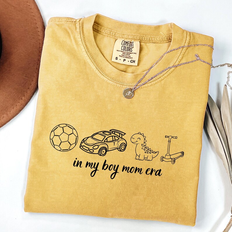 May include: Mustard yellow Comfort Colors t-shirt with black graphic illustrations of a soccer ball, race car, dinosaur, and scooter. The text "in my boy mom era" is below the graphics. A silver necklace is draped across the shirt.