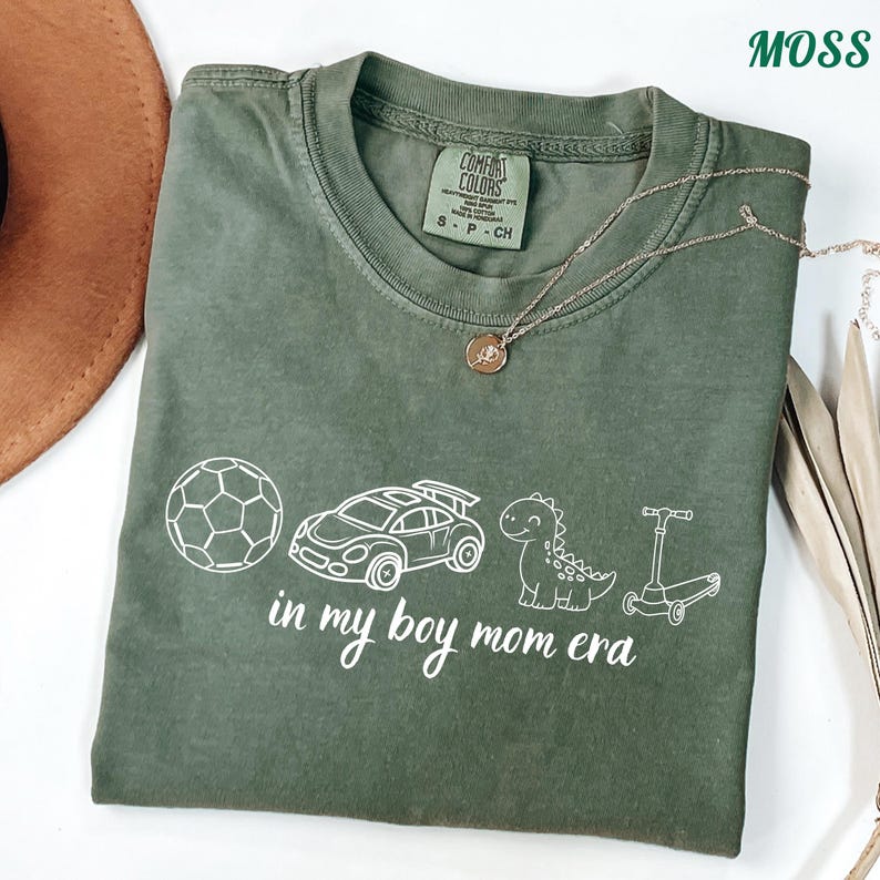 May include: A moss green t-shirt with the text "in my boy mom era" and white illustrations of a soccer ball, race car, dinosaur, and scooter. A gold necklace is draped across the shirt.