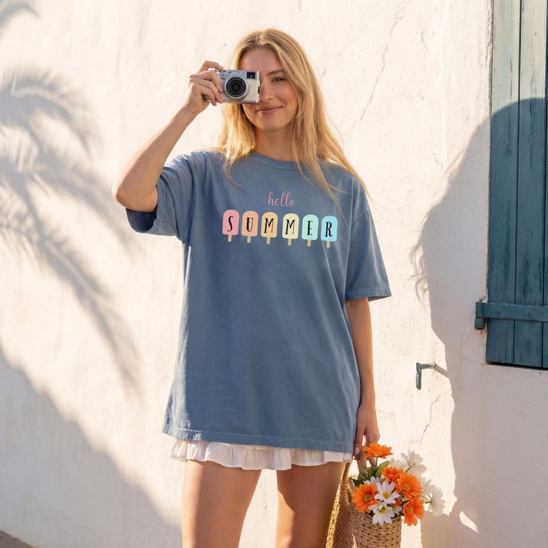 May include: A blue t-shirt with the words "hello SUMMER" printed in pastel colors. The shirt features a popsicle design. The person is holding a camera and wearing a white skirt. A straw bag with flowers is in the foreground.