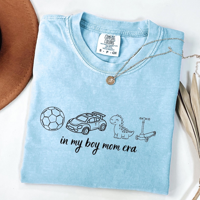 May include: Light blue t-shirt with a graphic of a soccer ball, race car, dinosaur, and scooter. The text "in my boy mom era" is printed below the graphics. A gold necklace is draped across the shirt.