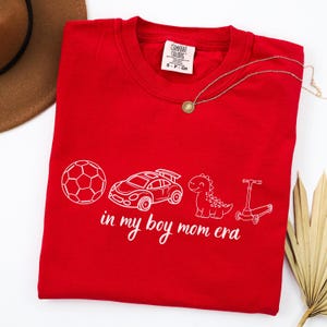 May include: A red Comfort Colors t-shirt with a white graphic of a soccer ball, race car, dinosaur, and scooter. The text "in my boy mom era" is below the graphics. A gold necklace is draped across the shirt.