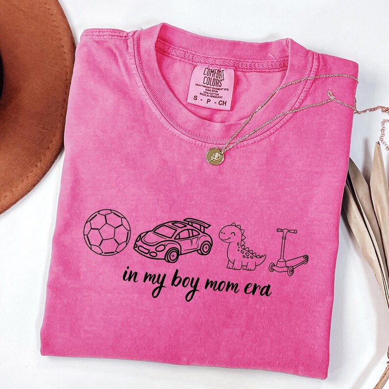 May include: A pink Comfort Colors t-shirt with a black graphic of a soccer ball, race car, dinosaur, and scooter. The text "in my boy mom era" is below the graphics. A gold necklace is draped across the shirt.