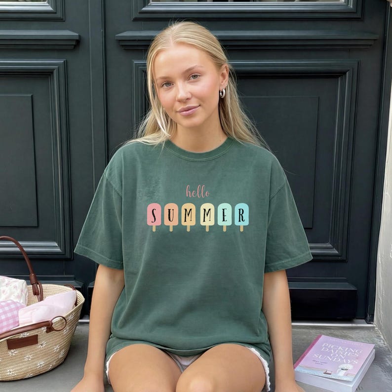 May include: Dark green t-shirt with the words "hello SUMMER" printed on the front. The word "hello" is in a cursive font, and "SUMMER" is in a block font with colorful popsicle graphics. The shirt is a casual, oversized style.