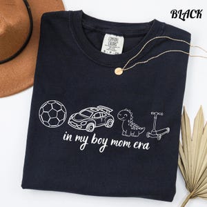 May include: Black t-shirt with white graphic of a soccer ball, race car, dinosaur, and scooter. The text "in my boy mom era" is below the graphics. A gold necklace and a brown hat are in the background.
