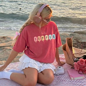 May include: A coral-colored t-shirt with the words "hello SUMMER" printed on the front in colorful popsicle-shaped letters. The shirt is paired with a white ruffled skirt. Accessories include pink headphones, sunglasses, and a straw tote bag.