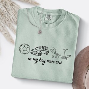 May include: A light green Comfort Colors t-shirt with a black graphic of a soccer ball, race car, dinosaur, and scooter. The text "in my boy mom era" is below the graphics. A silver necklace with a pendant is on the shirt.