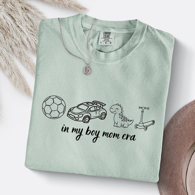 May include: A light green Comfort Colors t-shirt with a black graphic of a soccer ball, race car, dinosaur, and scooter. The text "in my boy mom era" is below the graphics. A silver necklace with a pendant is on the shirt.