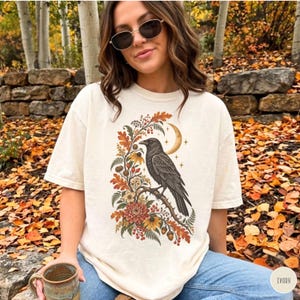 Könnte beinhalten: Ein elfenbeinfarbenes Kurzarm-T-Shirt mit einem Grafikdruck eines schwarzen Raben, der auf einem Ast sitzt, umgeben von Herbstlaub, Beeren, Blumen und einer Mondsichel mit kleinen Sternen, die mittig auf der Vorderseite platziert ist.