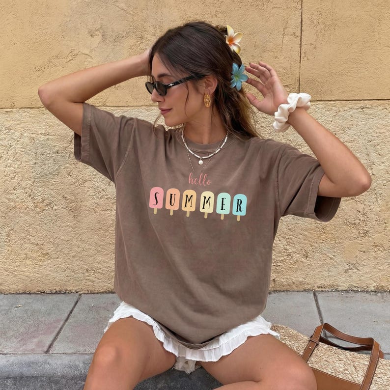 May include: A brown t-shirt with the words "hello SUMMER" printed in pastel colors. The design features popsicle illustrations. The shirt is worn by a person wearing sunglasses and a white scrunchie.