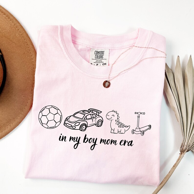 May include: A light pink t-shirt with a black graphic of a soccer ball, race car, dinosaur, and scooter. The text "in my boy mom era" is below the graphics. A rose gold necklace is draped on the shirt.