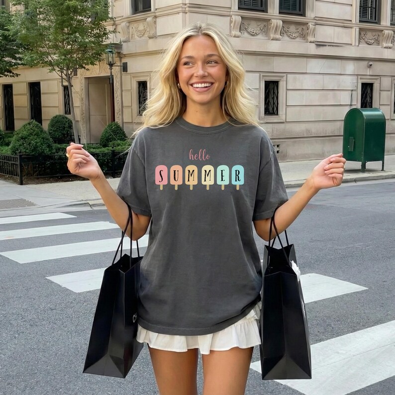 May include: Dark gray t-shirt with the words "hello SUMMER" above colorful popsicle graphics. The shirt is paired with a white skirt and black shopping bags. The image is taken outdoors.