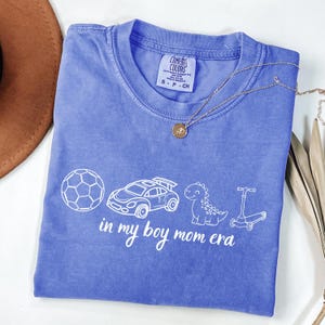 May include: A periwinkle blue t-shirt with the text "in my boy mom era" and illustrations of a soccer ball, race car, dinosaur, and scooter. A gold necklace with a pendant is on the shirt.