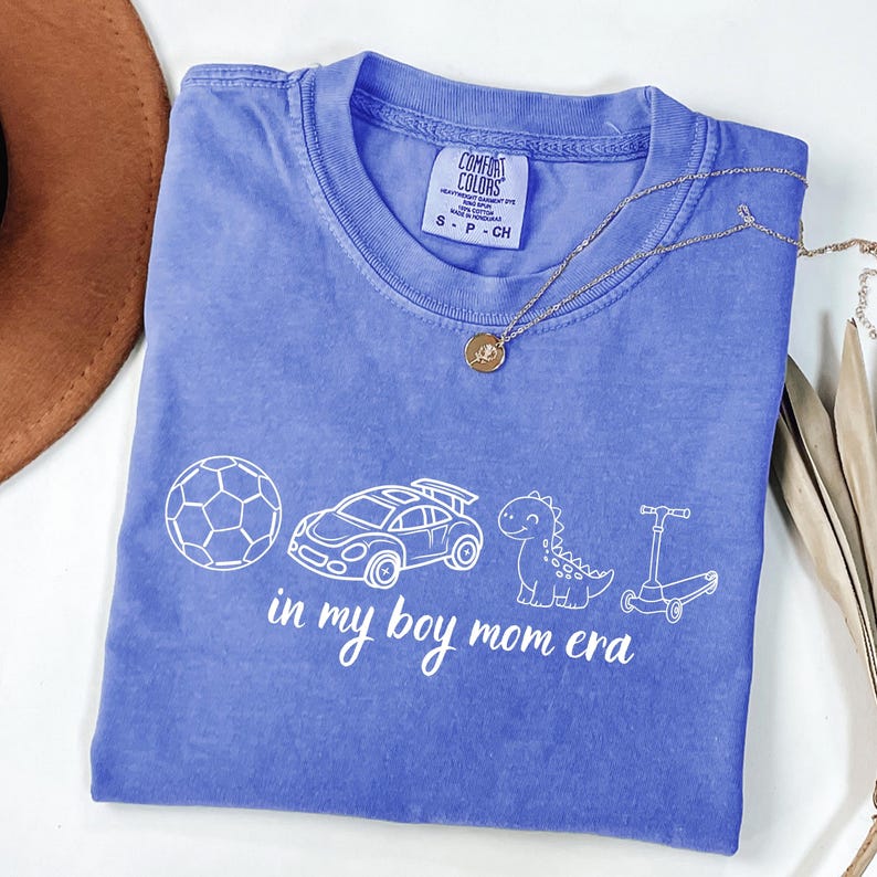 May include: A periwinkle blue t-shirt with the text "in my boy mom era" and illustrations of a soccer ball, race car, dinosaur, and scooter. A gold necklace with a pendant is on the shirt.
