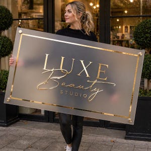 Peut inclure: Panneau rectangulaire avec l'inscription "LUXE Beauty STUDIO" en lettres dorées. Le panneau a une bordure dorée et est tenu par une personne. Le panneau est givré et a un design moderne.