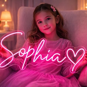 May include: A neon sign spelling "Sophia" in pink, with a heart design. The sign is held up. The background features soft lighting and a person wearing a pink dress. The sign is a decorative item.