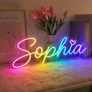 Può includere: Un'insegna al neon a forma del nome "Sophia" in corsivo, con un piccolo cuore sopra la "i". L'insegna si illumina con un gradiente arcobaleno di colori. L'insegna è posizionata su una superficie bianca e soffice.