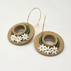 Könnte beinhalten: Ein Paar Ohrringe mit Miniatur-Strohhüten mit weißen Blumenakzenten und grünen Verzierungen. Die Ohrringe haben goldfarbene Creolenverschlüsse, und die Hüte sind beige mit einer gewebten Textur.