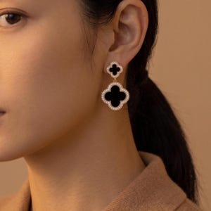 Peut inclure: Boucles d'oreilles pendantes élégantes avec un motif de trèfle à quatre feuilles. Les boucles d'oreilles ont un centre noir entouré d'un halo de cristaux étincelants. Le trèfle supérieur est plus petit que celui du bas. En métal doré.