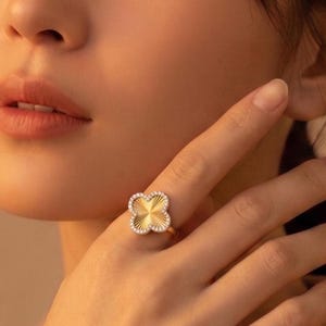 May include: A gold ring featuring a four-leaf clover motif with a sunburst texture at the center, surrounded by a border of small, clear, sparkling stones. The clover shape is symmetrical with rounded edges, set on a thin, polished gold band.