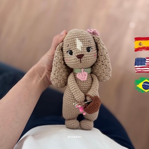 Pode incluir: Um cão de pelúcia feito em crochê na cor marrom claro com orelhas longas e caídas, uma mancha branca na testa, uma coleira verde clara com um pingente de coração rosa e uma pequena cesta marrom com flores rosas e brancas nas patas.