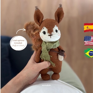 Puede incluir: Un juguete de ardilla tejido a ganchillo en marrón y blanco con una bufanda verde y un pequeño accesorio de bellota marrón. La ardilla tiene grandes ojos negros y orejas puntiagudas. Un gráfico circular blanco con una flecha indica 2 tail options included: fluffy or classic.