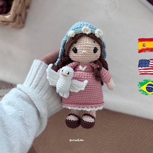 Puede incluir: Muñeca de ganchillo hecha a mano, con cabello castaño, velo azul y vestido rosa. La muñeca sostiene una paloma blanca. Los zapatos de la muñeca son marrones y blancos. La muñeca es una figura religiosa.