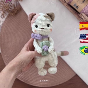 Puede incluir: Un muñeco de gato tejido a ganchillo hecho a mano con un cuerpo color crema y manchas de color rosa claro y marrón en las orejas. El gato lleva un lazo lavanda alrededor del cuello y sostiene un pequeño objeto cilíndrico verde con una flor.