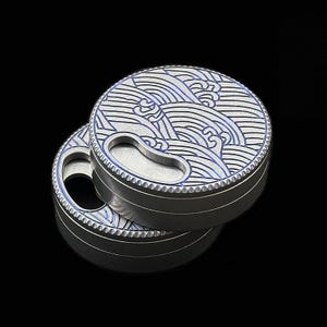 May include: Two round, silver-colored containers with a wave pattern in blue and silver. The lids have a keyhole-shaped opening. The containers are stacked on top of each other against a black background.